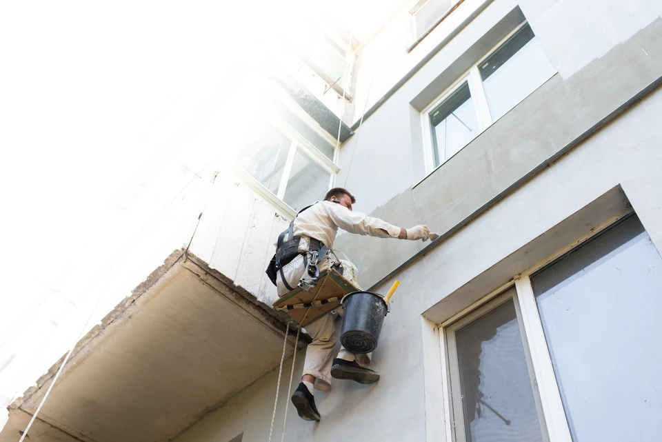 Ouvrier en tenue blanche effectuant des travaux de ravalement sur façade extérieure d'immeuble, suspendu par harnais de sécurité