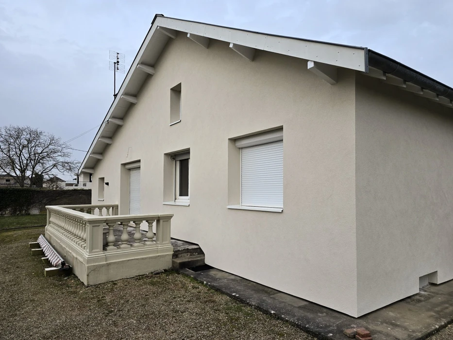 Façade latérale d'une maison individuelle rénovée, enduit de façade ton beige clair, volets roulants PVC blanc, fenêtres double vitrage à dormant PVC