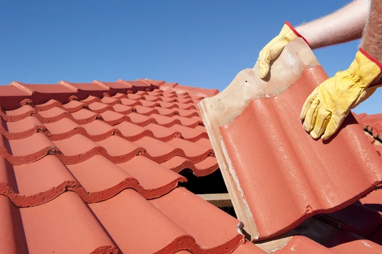 Ouvrier en gants de protection jaunes installant des tuiles sur une toiture en pente sous un ciel bleu dégagé