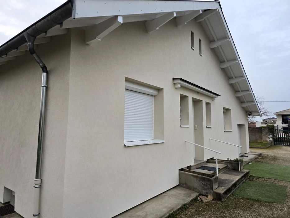Façade extérieure d'une maison individuelle plain-pied, enduit monocouche beige, volet roulant PVC blanc sur fenêtre à appui, porte d'entrée avec auvent tuiles canal, rampe d'accès PMR acier blanc