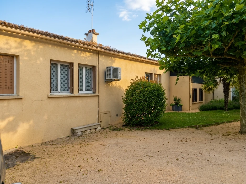 Façade latérale d'une maison individuelle rénovée, enduit minéral ton beige clair, volets roulants PVC blanc, fenêtres double vitrage, charpente apparente bois peint gris, débord de toit avec gouttière zinc