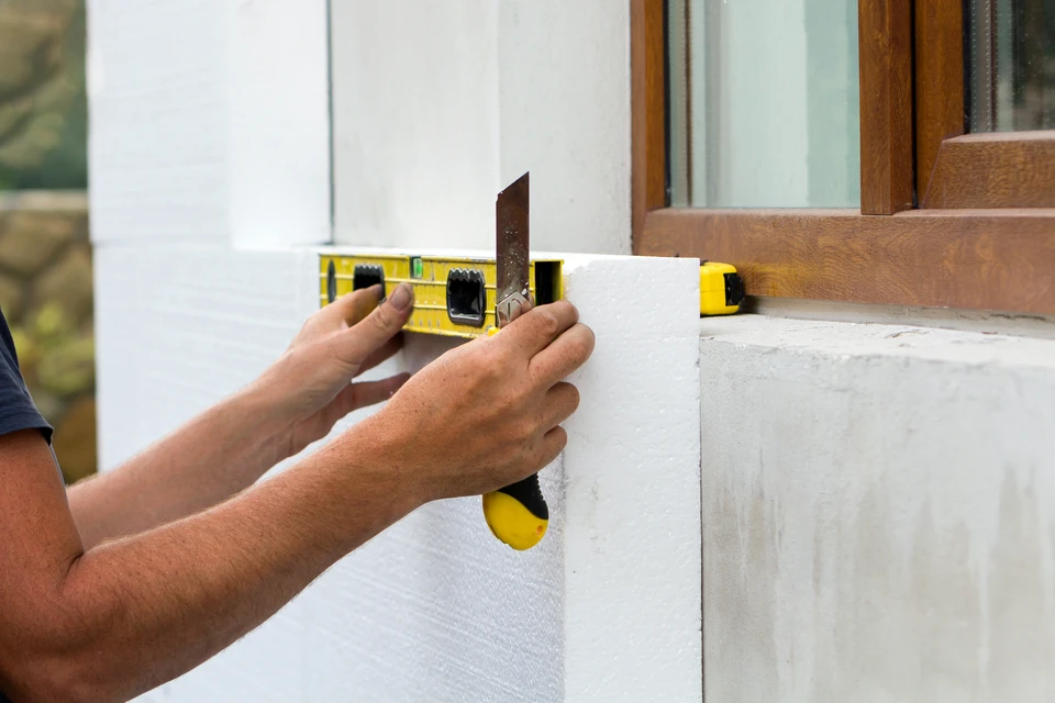 Chantier extérieur de rénovation de façade avec ouvrier en combinaison blanche appliquant un enduit sur mur beige