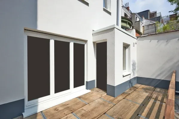 Vue en plongée légère d'un ouvrier qualifié en tenue professionnelle travaillant sur l'échafaudage d'une maison résidentielle. L'ouvrier, équipé de matériel professionnel, effectue le ravalement de la façade : nettoyage haute pression ou brossage de la surface, suivi de l'application de peinture ou d'enduit de finition. La façade du bâtiment est partiellement traitée, montrant le contraste entre les zones rénovées (propres et peintes) et les zones en cours de traitement. L'environnement montre un chantier professionnel de rénovation résidentielle, avec équipements et matériaux visibles. La lumière naturelle met en valeur le travail en hauteur et le professionnalisme de l'intervention.